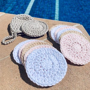 May include: A collection of circular, hand-crocheted pads in shades of white, light pink, beige, and gray. One gray pad features a small loop attached to its edge. The pads are arranged in a stacked, overlapping formation on a textured surface.