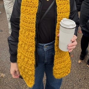 May include: A chunky mustard yellow knit scarf worn over a black jacket and black shirt, paired with dark blue denim jeans. A white travel mug with a lid is held in one hand, and a thin silver necklace with a small round pendant is visible.