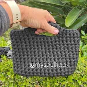 May include: A dark charcoal grey handbag crafted with a thick, chunky crochet stitch pattern. The bag features integrated handles and displays the white text @mysoulhook printed directly on the front surface of the textured, woven fabric.