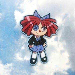 Op de afbeelding: Een sticker met een cartoonpop met rood haar, een blauwe strik en een zwart jasje over een blauwe rok. De pop heeft een knoogje als oog en lacht. De sticker heeft een lichtblauwe rand en een hemelachtergrond.