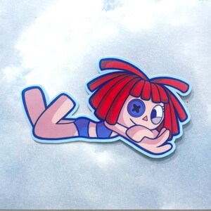 Op de afbeelding: Een sticker met een cartoonpop met rood haar, een knoogje en een blauw badpak. De pop ligt met uitgestrekte armen op een lichtblauwe achtergrond.