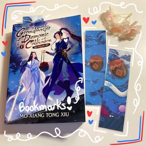 May include: A book and two bookmarks featuring artwork from "Grandmaster of Demonic Cultivation." The book cover shows two figures in flowing robes. The bookmarks have a blue background with illustrations of jars and landscapes. The word "Bookmarks" is written on the book.