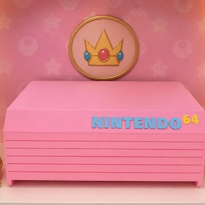 以下が含まれることがあります： ピンク色のNintendo 64コンソール。青と黄色の「NINTENDO 64」の文字。王冠、ピーチ姫の額入りアート、ハート型の装飾品など、テーマ別の装飾に囲まれています。