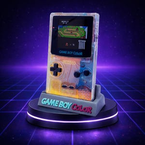 May include: A transparent Game Boy Color handheld console, displaying a retro video game on its screen. The console is on a gray stand with the words "GAME BOY COLOR" in teal and red. The background is a purple and blue grid.