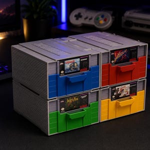 May include: A set of four gray plastic storage drawers designed to hold video game cartridges. Each drawer features a colorful front panel in blue, red, green, or yellow, with a visible game cartridge inserted into the top slot of each unit.
