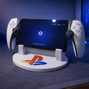 May include: A white gaming console stand featuring the PlayStation logo in red, yellow, and blue. The stand supports a black tablet with white controllers. The tablet screen displays the PlayStation logo on a blue background.