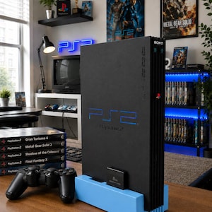 May include: A black PlayStation 2 console with a blue stand, controller, and game cases. The console has the PlayStation 2 logo in blue. Game titles include Gran Turismo 4, Metal Gear Solid 2, and Final Fantasy X.