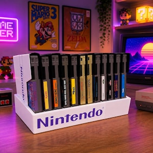 May include: A white plastic display stand featuring the Nintendo logo in purple text, holding a collection of twelve gray and gold rectangular video game cartridges. The stand is designed to organize and showcase classic gaming cartridges.