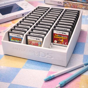 May include: A white Nintendo DS game cartridge organiser filled with game cartridges. The organiser is on a colourful chequered surface, with a Nintendo DS console and game cartridges in the background. Two blue styluses are in the foreground.