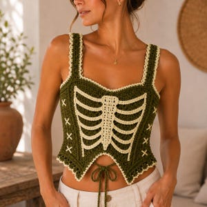 May include: A dark olive green crochet tank top featuring a cream-colored rib cage design on the front. The garment has thick straps, a square neckline, and a pointed hemline with a central tie closure. The edges are finished with a cream trim.