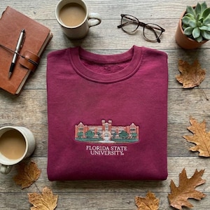 Puede incluir: Una sudadera granate con una imagen bordada de un edificio universitario y el texto "FLORIDA STATE UNIVERSITY." La sudadera está doblada sobre una superficie de madera, rodeada de tazas de café, un diario, gafas y hojas de otoño.