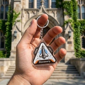May include: A clear acrylic keychain featuring a triangular design with a space shuttle illustration. The keychain has the text "1984" and "2011" on the sides, and "Discovery" at the bottom. The keychain is held in a hand.