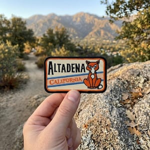 May include: Rectangular patch with the text "ALTADENA CALIFORNIA" and a cartoon fox illustration. The patch has a black border, with blue and orange stripes. The fox is orange and white, sitting upright.