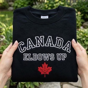 May include: Black sweatshirt with the word "CANADA" in white block letters, outlined in white, arched across the top. Below, the words "ELBOWS UP" are in white block letters. A red maple leaf is at the bottom. The sweatshirt is folded.