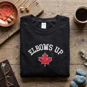 May include: Black crewneck sweatshirt with the text "ELBOWS UP" in white arched lettering above a red maple leaf. The sweatshirt is displayed on a wooden surface with a cup of coffee, a notebook, and a bowl with a red leaf.