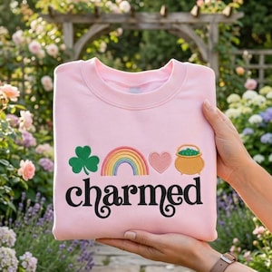 Könnte beinhalten: Ein hellrosa Sweatshirt mit dem Wort "charmed" in schwarzer Schrift. Über dem Wort befinden sich gestickte Bilder eines grünen Kleeblatts, eines Regenbogens, eines rosa Herzens und eines Goldtopfes. Das Sweatshirt wird von einer Person gehalten.
