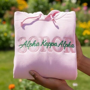 Puede incluir: Una sudadera con capucha rosa claro con el texto bordado "Alpha Kappa Alpha" en verde y rosa. La sudadera tiene un cordón y está sostenida por las manos de una persona. El fondo es un jardín borroso.