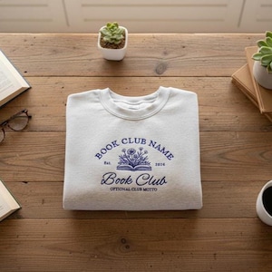 May include: A light gray sweatshirt with a crew neck, featuring a navy blue design that reads "BOOK CLUB NAME" above an open book illustration with flowers, and "Book Club" below. The text "Est. 2016" and "Optional Club Motto" are also included.