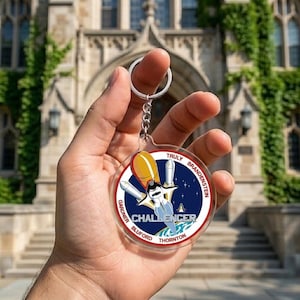 May include: Clear acrylic keychain featuring a circular design with a rocket ship and the word "CHALLENGER". The design includes the names of astronauts and a blue and white color scheme. The keychain has a silver chain and ring.