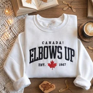 May include: White sweatshirt with the text "CANADA! ELBOWS UP EST. 1867" in black lettering and a red maple leaf. The sweatshirt is folded on a wooden surface, with a book and coffee cup in the background.