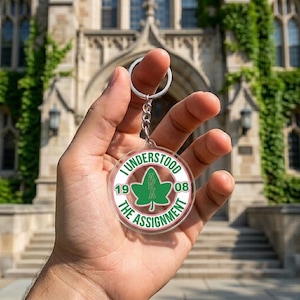 May include: Clear acrylic keychain with a silver chain and ring. The keychain features a green and white design with the text "I UNDERSTOOD THE ASSIGNMENT" and "AKA 1908". The background is a building with ivy.