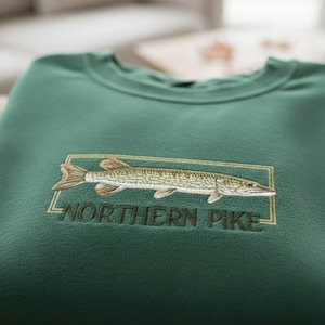 May include: A forest green sweatshirt featuring an embroidered design of a Northern Pike fish within a rectangular frame. The text "NORTHERN PIKE" is embroidered below the fish. The sweatshirt is made of a soft, textured fabric.