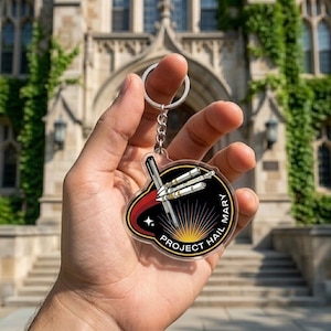 May include: A clear acrylic keychain with a silver keyring and chain. The keychain features a black and red emblem with the text "PROJECT HAIL MARY" and a graphic of three rockets. The background is a blurred image of a building.