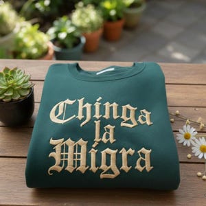 Könnte beinhalten: Ein gefaltetes, dunkelgrünes Sweatshirt mit dem gestickten Text "Chinga la Migra" in goldener, gotischer Schrift. Das Sweatshirt liegt auf einer Holzoberfläche, mit einer Sukkulente und Gänseblümchen im Hintergrund.