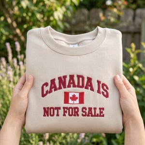 May include: A beige sweatshirt with the words "CANADA IS NOT FOR SALE" embroidered in red. The design includes a red and white Canadian flag. The sweatshirt is folded and held by hands.
