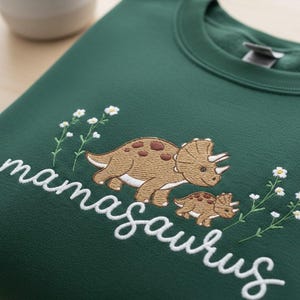 May include: Dark green sweatshirt with embroidered text reading "mamasaurus" and a dinosaur design. The design features a brown triceratops with a baby triceratops and white flower accents.