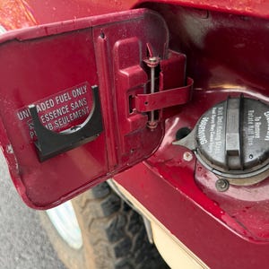 May include: A close-up of a red vehicle's open fuel door. The door has a black and white sticker with text in English and French. The fuel cap is visible, with a warning label. The vehicle's tire is partially visible.