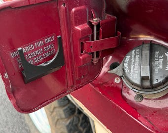 Puede incluir: Primer plano de la puerta de combustible abierta de un vehículo rojo. La puerta tiene una pegatina en blanco y negro con texto en inglés y francés. La tapa del combustible es visible, con una etiqueta de advertencia. El neumático del vehículo es parcialmente visible.