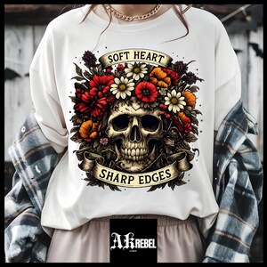 May include: White t-shirt featuring a skull design adorned with red, orange, and white flowers. The design includes banners with the phrases "SOFT HEART" and "SHARP EDGES". The shirt has a crew neck.