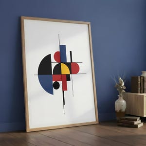 May include: A framed abstract art print featuring geometric shapes in red, black, yellow, and blue. The artwork is set against a white background and framed in natural wood. The print leans against a blue wall.