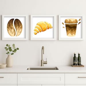 Könnte beinhalten: Drei quadratische weiße Rahmen mit Illustrationen einer Kaffeebohne, eines goldenen Croissants und einer Kaffeetasse mit Spritzern. Die Kunstwerke zeigen Braun-, Beige- und Gelbtöne auf weißem Hintergrund und hängen in einer Küche.