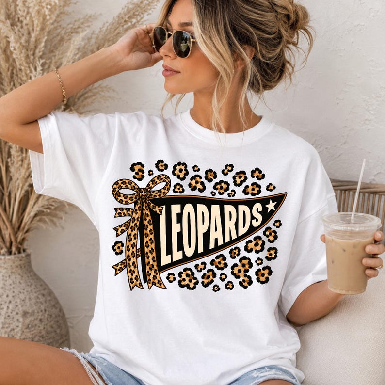 May include: A white short-sleeved t-shirt featuring a graphic of a black pennant flag with the word LEOPARDS in cream-colored letters. The design includes a large leopard-print bow and scattered leopard-print spots surrounding the pennant.