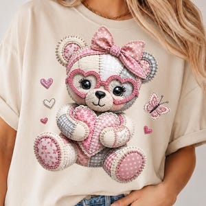 Può includere: Una t-shirt beige con la grafica di un orsacchiotto patchwork con un fiocco rosa, occhiali a forma di cuore e una toppa a forma di cuore sul petto. L'orso è circondato da piccoli cuori rosa e una farfalla, il tutto in stile cucito.