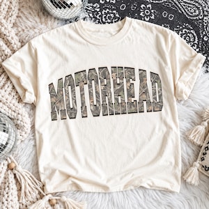 Puede incluir: Una camiseta de manga corta de color crema con la palabra MOTORHEAD impresa en una fuente grande y arqueada de estilo universitario. Las letras están rellenas con un patrón de camuflaje forestal realista en tonos verde, marrón y beige.