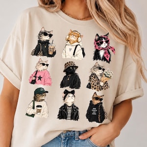 Può includere: Una t-shirt beige a maniche corte con una stampa grafica di nove gatti stilizzati vestiti con vari abiti, tra cui occhiali da sole, cappelli, sciarpe e felpe con cappuccio. Un gatto indossa una felpa con la scritta More Espresso Less Drama.