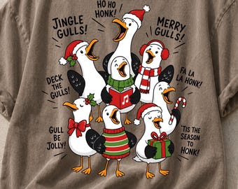 May include: A brown t-shirt featuring a graphic of seven seagulls wearing holiday accessories like Santa hats, scarves, and sweaters. The text reads: JINGLE GULLS!, HO HO HONK!, MERRY GULLS!, DECK THE GULLS!, FA LA LA HONK!, GULL BE JOLLY!, 'TIS THE SEASON TO HONK!