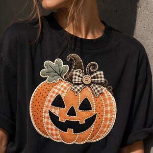 May include: A black t-shirt featuring a large, central appliqué of a jack-o'-lantern pumpkin. The pumpkin is crafted from orange fabric with polka dot and gingham patterns, a green leaf, and a black and white chequered bow with a wooden button.