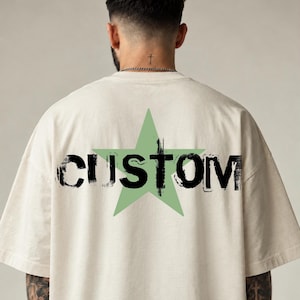 May include: An off-white short-sleeve t-shirt featuring a large, light green five-pointed star graphic on the back. The word CUSTOM is printed across the centre of the star in a bold, black, distressed, and textured font style.