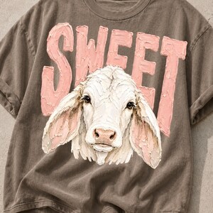 May include: A brown short-sleeved t-shirt featuring a graphic of a white cow head with large ears. Behind the cow, the word SWEET is printed in large, textured, light pink capital letters. The design has a thick, impasto-style paint appearance.