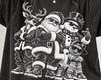 May include: A black t-shirt featuring a graphic print of a reindeer, Santa Claus, a snowman, and a gingerbread person, all wearing sunglasses. The characters are dressed in winter attire and are standing in a group with their arms crossed.
