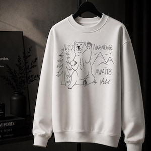 May include: A white crewneck sweatshirt featuring a black line art graphic of a bear waving, a pine tree, a sun, and mountains. The text "ADVENTURE AWAITS" is printed in a simple, handwritten-style font next to the bear illustration.