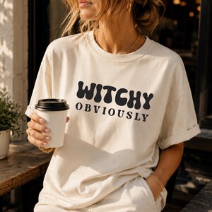 May include: A cream-colored short-sleeve t-shirt featuring the black text WITCHY OBVIOUSLY printed on the front in a bold, wavy font. The shirt has a crew neckline and a relaxed fit, with the sleeves slightly rolled at the edges for a casual look.