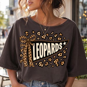 May include: A charcoal gray short-sleeve t-shirt featuring a graphic print of a black pennant flag with the word LEOPARDS in white capital letters. The design includes a leopard print bow and leopard print spots surrounding the pennant.