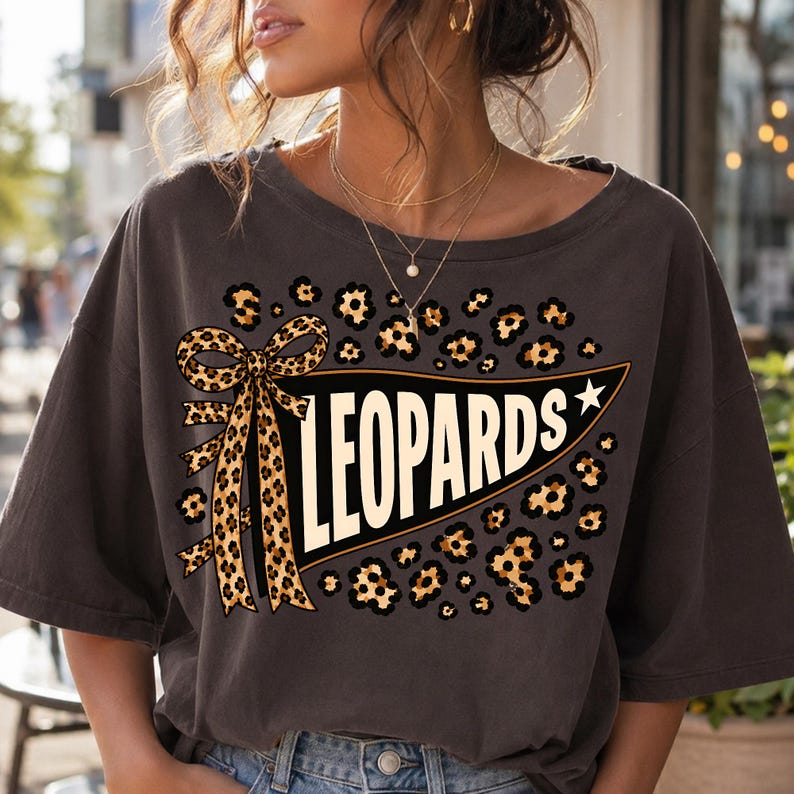 May include: A charcoal gray short-sleeve t-shirt featuring a graphic print of a black pennant flag with the word LEOPARDS in white capital letters. The design includes a leopard print bow and leopard print spots surrounding the pennant.