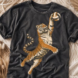May include: A charcoal grey acid-wash t-shirt featuring a graphic print of an orange and black tiger reaching upward to strike a volleyball. The tiger is depicted in a dynamic jumping pose with its front paw extended toward the ball.