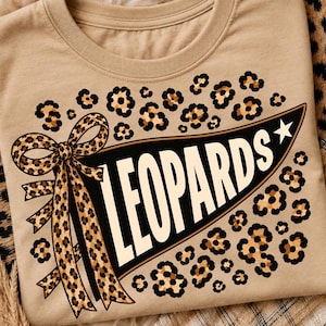 May include: A tan t-shirt featuring a black pennant graphic with the word LEOPARDS in white capital letters. The design includes a leopard print bow on the left side of the pennant, a white star on the right, and leopard print spots scattered around.